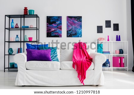 Vertical view of stylish living room with comfortable white couch with pink blanket and blue and purple pillows, galaxy graphics on the wall and metal shelves with accessories, real photo