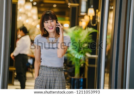 A portrait of a middle-aged, classy and elegant Asian Chinese woman talking on her smartphone as she stands in the doorway indoors. She is smiling as she talks animatedly.