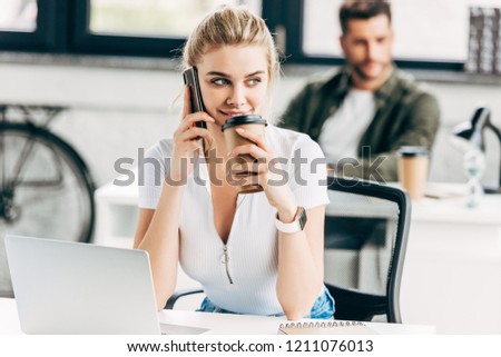 beautiful young woman talking by phone and drinking coffee at office with colleague on background