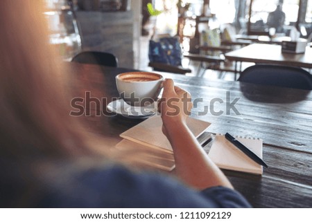 Closeup image of a woman holding and drinking hot coffee with notebooks on table in cafe