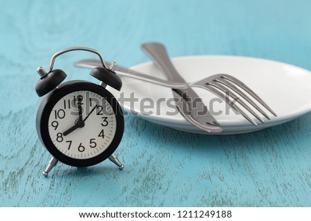 Clock with white plate, fork and knife, intermittent fasting, diet, weight loss concept on blue wooden table Royalty-Free Stock Photo #1211249188