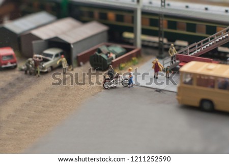 a miniature depicting a Parking lot at the station where the biker flirts with the beauty, and next to people go to the bus standing next to the background of the garages in which men are busy with th