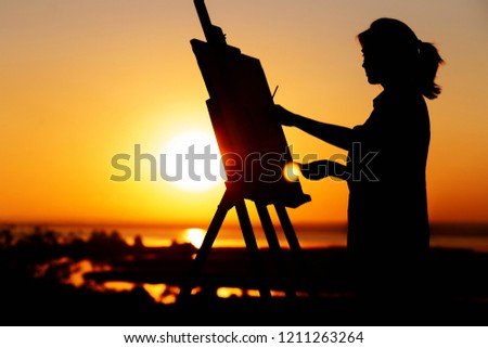 silhouette of a young woman painting a picture with paints on canvas on an easel outdoors, girl with paint brush and palette engaged in art on the nature in a field at sunset