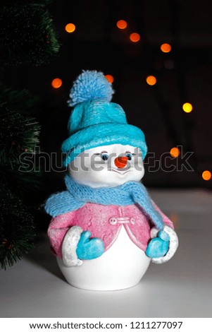  Toy snowman in pink jacket, blue hat, scarf and mittens near Christmas tree on dark background with multicolor lights Christmas  bokeh 