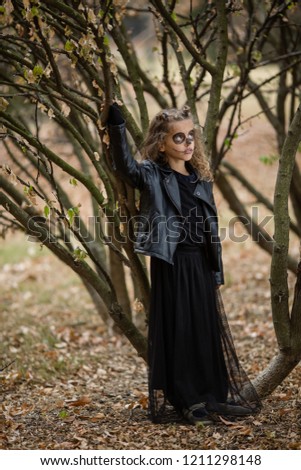 Cute little goth girl dressed in black leather jackets, painted face with scary crayonne drawings. Halloween costume. Spooky tree in background. Blond, curly hair. Rock and roll kids. Heavy metal punk