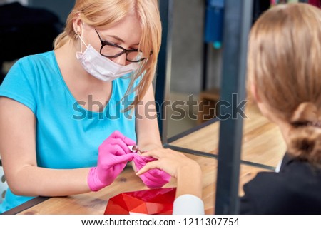 Qualified beauty specialist in process of doing manicure to woman sitting opposite in salon. Worker in protective gloves and mask covering nails with brown varnish. Concept of beauty procedure.