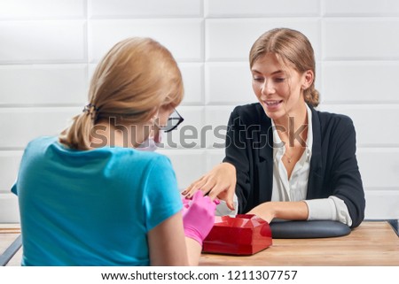 Beautiful woman in office suit talking with manicurist sitting opposite while she doing professional manicure in salon. Beauty specialist in protective gloves and mask covering nails with varnish.