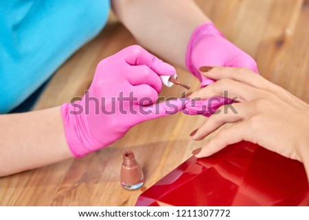 Closeup of process of covering nails with brown varnish to female customer in beauty salon. Skilled beautician in protective gloves doing professional manicure to woman. Concept of cosmetic procedure.