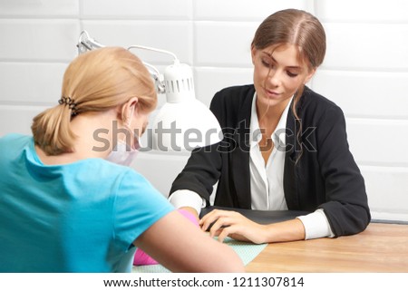 Attractive lady in office suit sitting at table, receiving manicure and enjoying procedure in beauty salon. Professional manicurist in protective gloves and mask serving female customer.