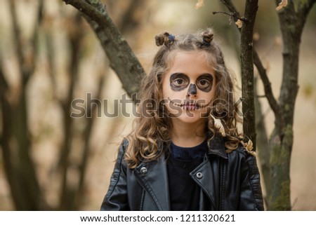 Cute little goth girl dressed in black leather jackets, painted face with scary crayon drawings. Halloween costume. Spooky tree in background. Blond, curly hair. Rock and roll kids. Looking sad, alone