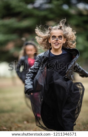 Two cute goth girls in black leather jackets, running behind one another with painted face,scary crayon drawings. Halloween costume. Nature in autumn. Blond, curly hair. Rock and roll kids. In motion.