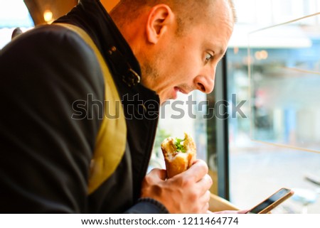 Digital lottery winner. A man eats a sandwich in a bakery next to a large window with natural light and uses a smartphone to do the job. Man Using Digital Device Concept. Surprise with news. 
