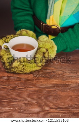 Women's hands in mittens holding a mug on the background of a knitted sweater, wooden background. Winter and Christmas concept.