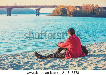 A single man tourist sitting on the river bank in autumn and looking at water 