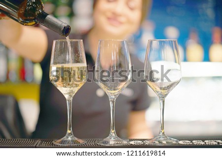 Bartender woman standing smiling, pours white wine into a glass from a bottle. Shelves with bottles of alcohol in the background. Focus on the glass.