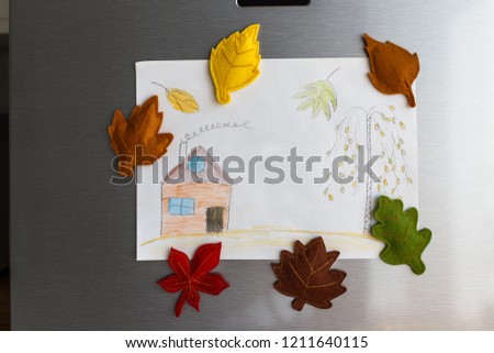 Colorful felt autumn leaves on refrigerator door. Children picture with house and tree hanging on fridge door. White kitchen on background. Diy fall autumn decoration for home