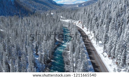 AERIAL: Flying above a breathtaking emerald river running along forest road. Picturesque aerial view of the winter landscape in the American wilderness. Vivid stream rushing through the countryside.