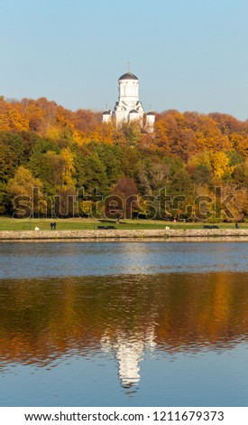 Colorful autumn. Alley along the riverbank on background of the autumn park. Reflections in the water of the forest and blue sky. Over all towers the Orthodox Church of Beheading of John the Baptist