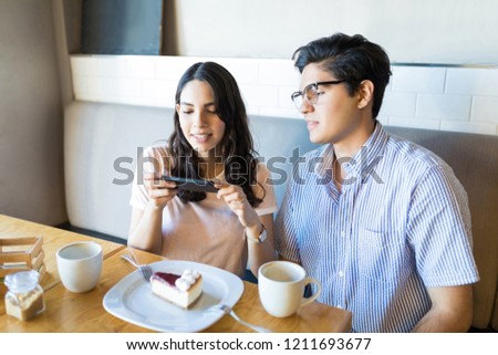 Young guy looking at girlfriend taking picture of cheesecake in cafe