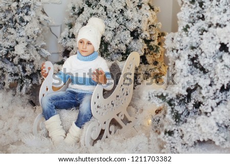 Boy in a white and blue sweater and white hat is sitting in a decorative sleigh near the Christmas tree.