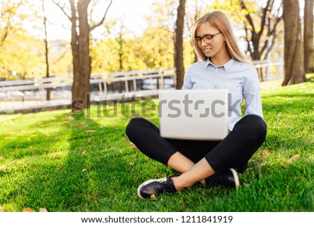 Young attractive girl with glasses, Woman sitting on the grass, working on a laptop, in a city park on a green lawn outdoors. Freelance business concept