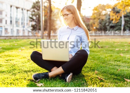 Portrait of pretty young woman in official clothes sitting on green grass in the park with legs crossed using a laptop