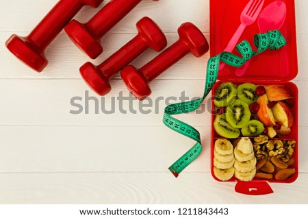  Kiwi, apple, bananas, nuts, dried fruits and momordica berries in a red box. Tape measure and dumbbells. The concept of a healthy lifestyle. Fitness menu. Banner. Top view. Flat lay