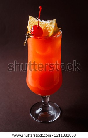 Glass of red alcohol cocktail with cherry, slice of orange and straw on elegant dark brown background.
