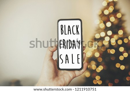Black Friday big sale text. Holiday discount offer. Hand holding phone on background of golden beautiful christmas tree with lights in festive room.