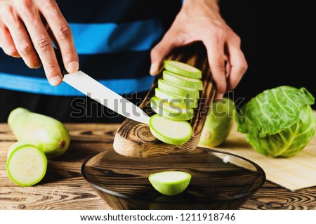 Man holds knife and pouring out pieces of fresh zucchini in glass bowl on old brown wooden table with kitchenware on black background
