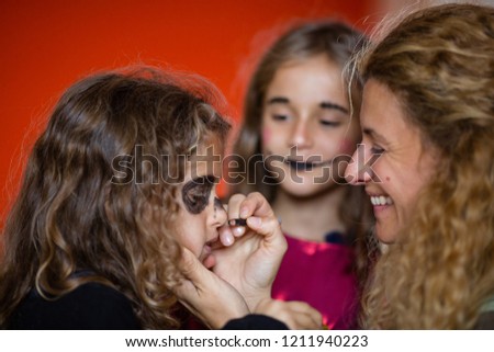 Blond curly young mother putting Halloween makeup on daughter face, other girl watching. Goth girl face paint. Orange blurred bokeh background. Children's fashion. Crayon drawings on face. Family fun.