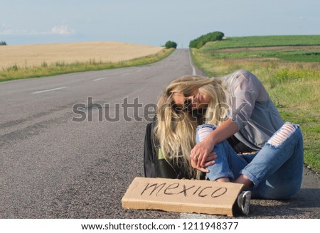 Serious blonde holding sign while hitchhiking on the road in summertime.