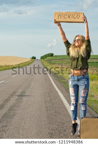 Blonde holding sign while hitchhiking on the road in summertime.