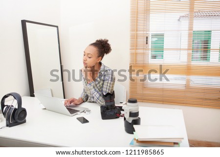 Portrait of beautiful african american media student girl using laptop computer and photographic equipment in a home desk office room, indoors. Telecommunication technology college studying home work.