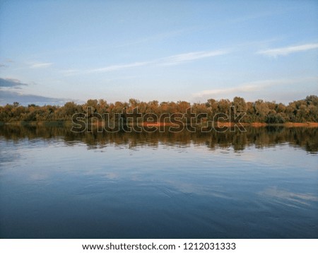 Evening panorama of the calm river bank with trees and clouds reflected in water.