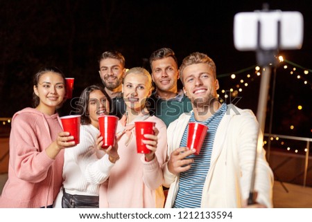 leisure, celebration and people concept - happy friends with drinks taking picture by selfie stick at rooftop party at night