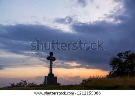 Silhouette of lonely orthodox cross on a hill covered with dried high grass with dramatic sunset sky background and the black birds