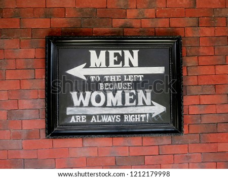 Graphite black board with the ironic inscription white chalk - "Men to the left because women are always right" on the  wall of red bricks. Humor, citation.