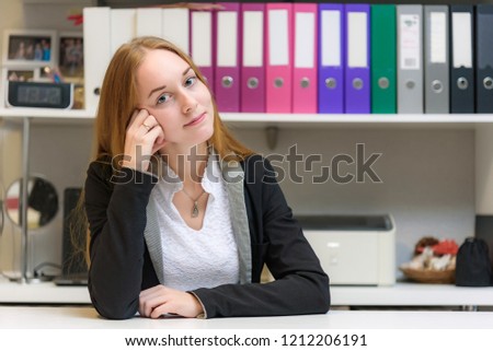 Concept manager in the office of a beautiful girl with long hair sitting at the table and talking. She is right in front of the camera smiling and looking happy.