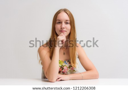 Studio portrait on camera of a smiling beautiful girl with long hair is talking sitting at a table with emotions. She stands right in front of the camera in various poses and looks happy.