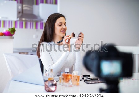 Young cute blogger woman filming her presentation of cosmetic products. Sitting in a bright room in front of camera.
