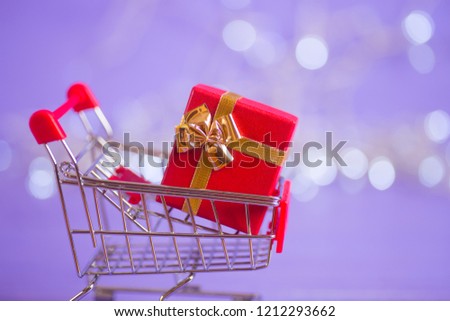Red gift box in small supermarket trolley with garland lights on purple bokeh background. New year banner with empty space. Concept of Valentine day shopping.
