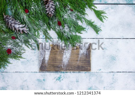 Blank wood sign with Christmas tree garland border, red berries and pine cones on antique rustic wooden snowy background; winter holiday sign with decorations and copy space