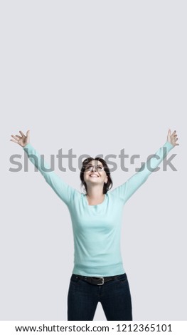 A happy brunette girl in a mint-colored jumper stands against the gray background The young woman raised her hands up  smiles broadly and looks up
