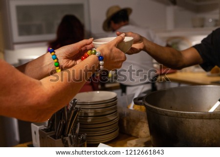 Woman with colorful beads bracelet is picking up hear soup bowl from the cooks - Low angle picture with a pile of bowls and many utensils in the background