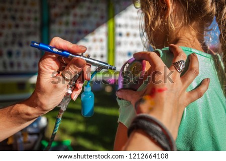 Young girl getting an airbrush stencil temporary tattoo in a family festival outdoors - Closeup picture with many different tattoo designs displayed in the blurry background Royalty-Free Stock Photo #1212664108