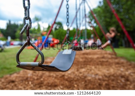 Closeup picture of a swing in a park for kids. Kids swigning in the blurry background - Picture taken on a warm summer day, with mulch ground instead of sand, and green lawn in the background