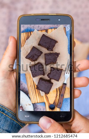 Smartphone photography of food. Woman hands holds mobile phone and take or make beautiful trendy food photo for social networks or blogging. Vegan chocolate brownie sweet cake for dessert