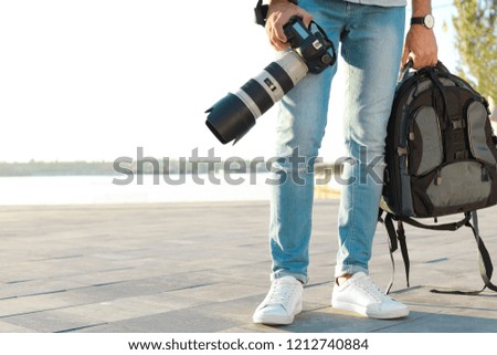 Young male photographer holding professional camera and backpack at pier. Space for text