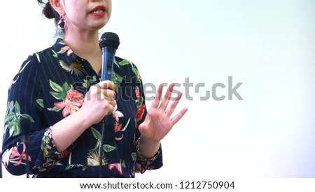 The woman is holding the microphone in  the left hand and do the public speaking on the white background.